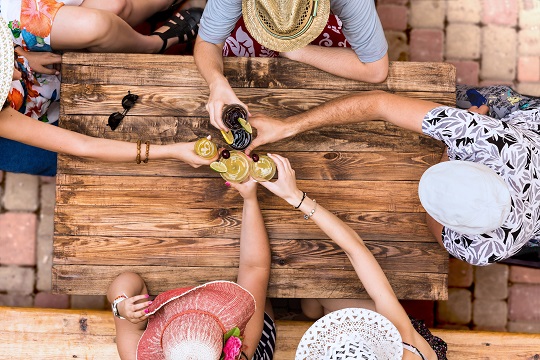 Hand of people cheers with alcohol cocktails at natural handcrafted wooden desk with brown sharp texture casual summer vacation style dress hats